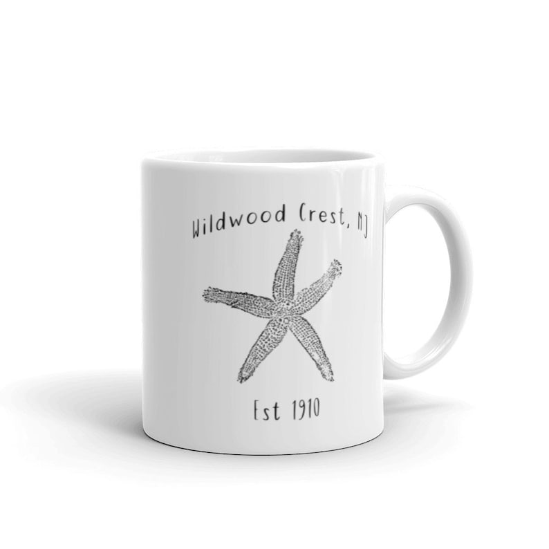 May include: White ceramic mug featuring a detailed black and white starfish design. The mug has the text "Wildwood Crest, NJ" above the starfish and "Est 1910" below it. The mug has a curved handle.