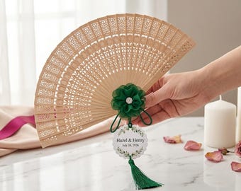 May include: A light brown wooden folding fan with intricate laser-cut patterns. The fan is decorated with a dark green fabric flower featuring a central rhinestone, and a white scalloped tag printed with "Hazel & Henry July 26, 2026" and a green tassel.