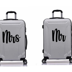 May include: Two silver suitcases with black handles and wheels. One suitcase has "Mrs" written on it in black cursive lettering. The other suitcase has "Mr" written on it in black cursive lettering.