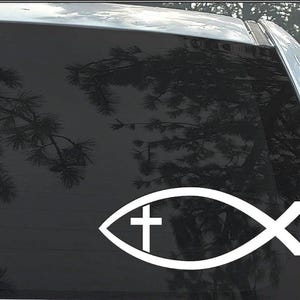May include: White vinyl decal of a fish with a cross inside. The decal is on a car window.
