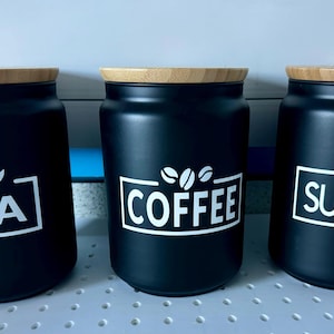 Può includere: Tre contenitori neri con coperchi in legno ed etichette bianche. Le etichette riportano "TEA", "COFFEE" e "SUGAR".