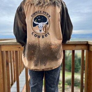 May include: A tan and brown button-up shirt with a graphic on the back that says "Simmer Down Cowboy" in black text. The graphic features a bull skull, cactus, and stars. The shirt is worn with gray jeans.