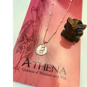 May include: A gold necklace with a round pendant featuring an owl design. The pendant is on a red background with the text "ATHENA Goddess of Wisdom and War".