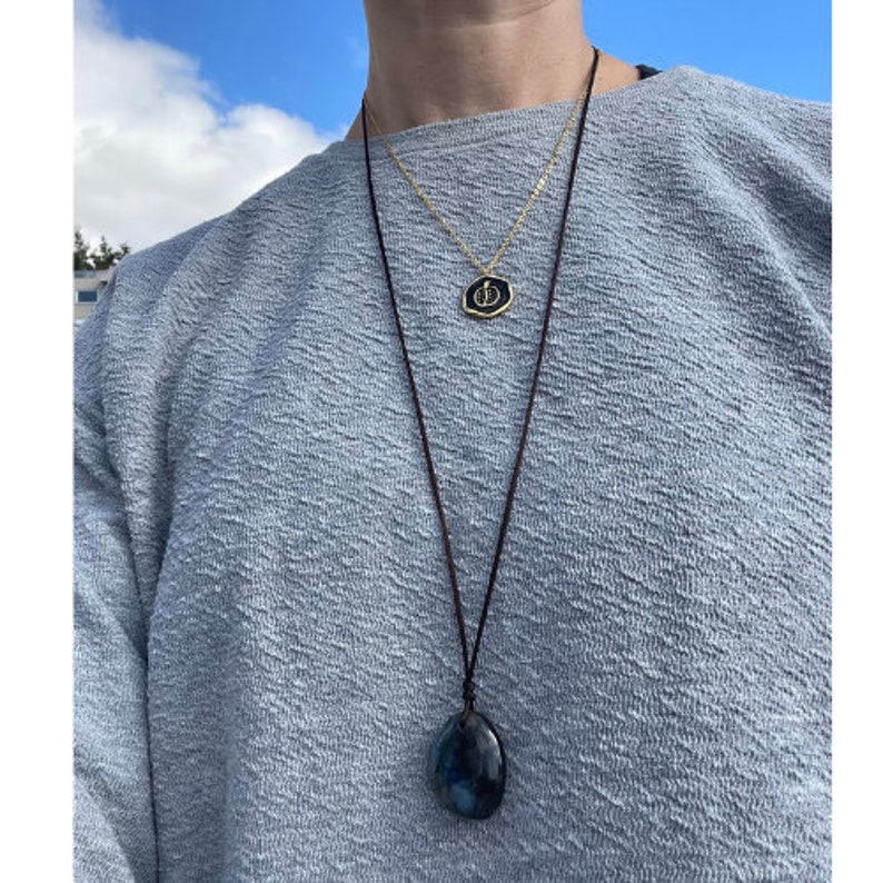 May include: A person wearing a light blue jumper with two necklaces. One necklace has a gold pendant with a black design. The other necklace has a dark blue stone pendant.