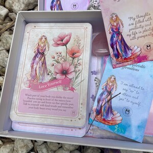 May include: A set of affirmation cards featuring a goddess with flowers and a quote about loving yourself. The cards are in a box with a pink and blue color scheme.