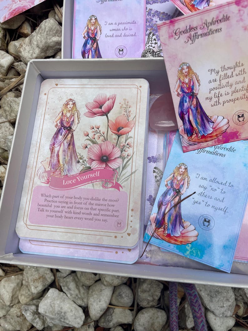 May include: A set of affirmation cards featuring a goddess with flowers and a quote about loving yourself. The cards are in a box with a pink and blue color scheme.