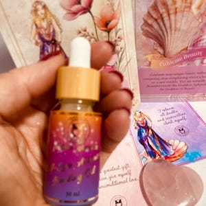 May include: A small glass bottle with a dropper filled with a clear liquid. The bottle has a gold label with the text "Rose Moon Facial Oil" and "30 ml". The bottle is sitting on a pink and white surface with a pink heart-shaped stone and a card with the text "Cultivate Beauty" and "I release all doubts and insecurities about myself".