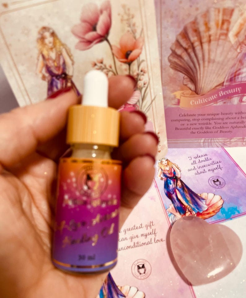 May include: A small glass bottle with a dropper filled with a clear liquid. The bottle has a gold label with the text "Rose Moon Facial Oil" and "30 ml". The bottle is sitting on a pink and white surface with a pink heart-shaped stone and a card with the text "Cultivate Beauty" and "I release all doubts and insecurities about myself".