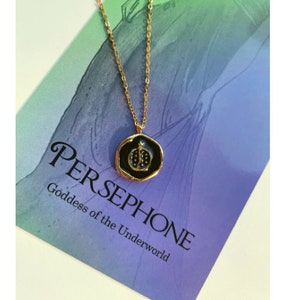May include: A gold chain necklace with a black enamel pendant featuring a pomegranate design. The pendant has the text "PERSEPHONE Goddess of the Underworld" on the card.