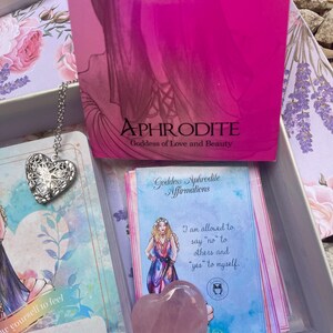 May include: A pink heart-shaped crystal with a blue and white card featuring an illustration of a woman and the text "Goddess Aphrodite Affirmations I am allowed to say "no" to others and "yes" to myself."