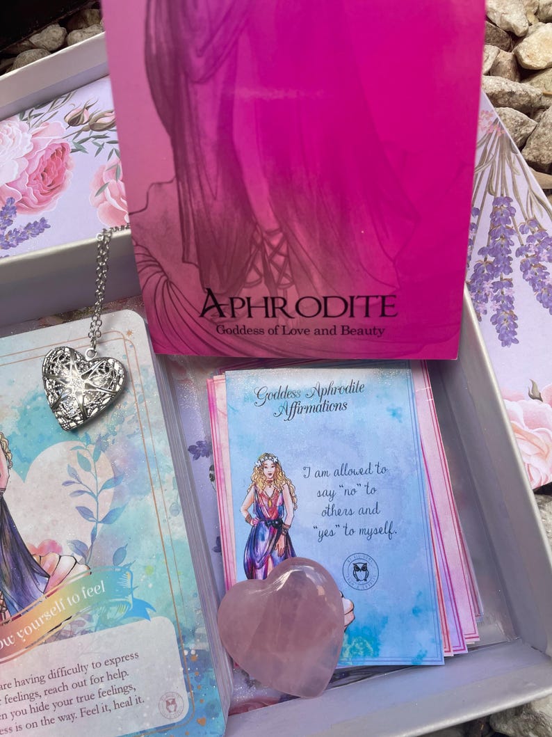 May include: A pink heart-shaped crystal with a blue and white card featuring an illustration of a woman and the text "Goddess Aphrodite Affirmations I am allowed to say "no" to others and "yes" to myself."
