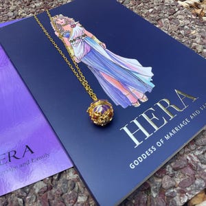 Puede incluir: Un collar dorado con un colgante redondo con una gema púrpura y detalles rojos, sobre un libro azul marino. El libro tiene una ilustración de una mujer con un vestido fluido y la palabra "HERA".