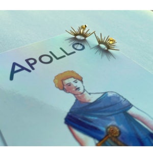 Könnte beinhalten: Ein Paar goldene Sonnenstrahlen-Ohrringe. Die Ohrringe sind klein und zart, mit einem schlichten Design. Die Ohrringe befinden sich auf einem weißen Hintergrund mit dem Wort "APOLLO" in Schwarz gedruckt.