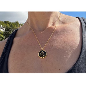 May include: A gold chain necklace with a black and gold pendant featuring a pomegranate design. The pendant is a hexagonal shape.