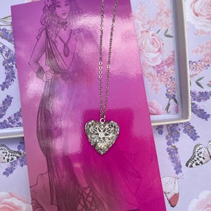 May include: A silver heart-shaped locket necklace with a delicate chain. The locket is adorned with intricate filigree designs and sparkling rhinestones. The necklace is displayed on a pink background with the text "APHRODITE Goddess of Love and Beauty".