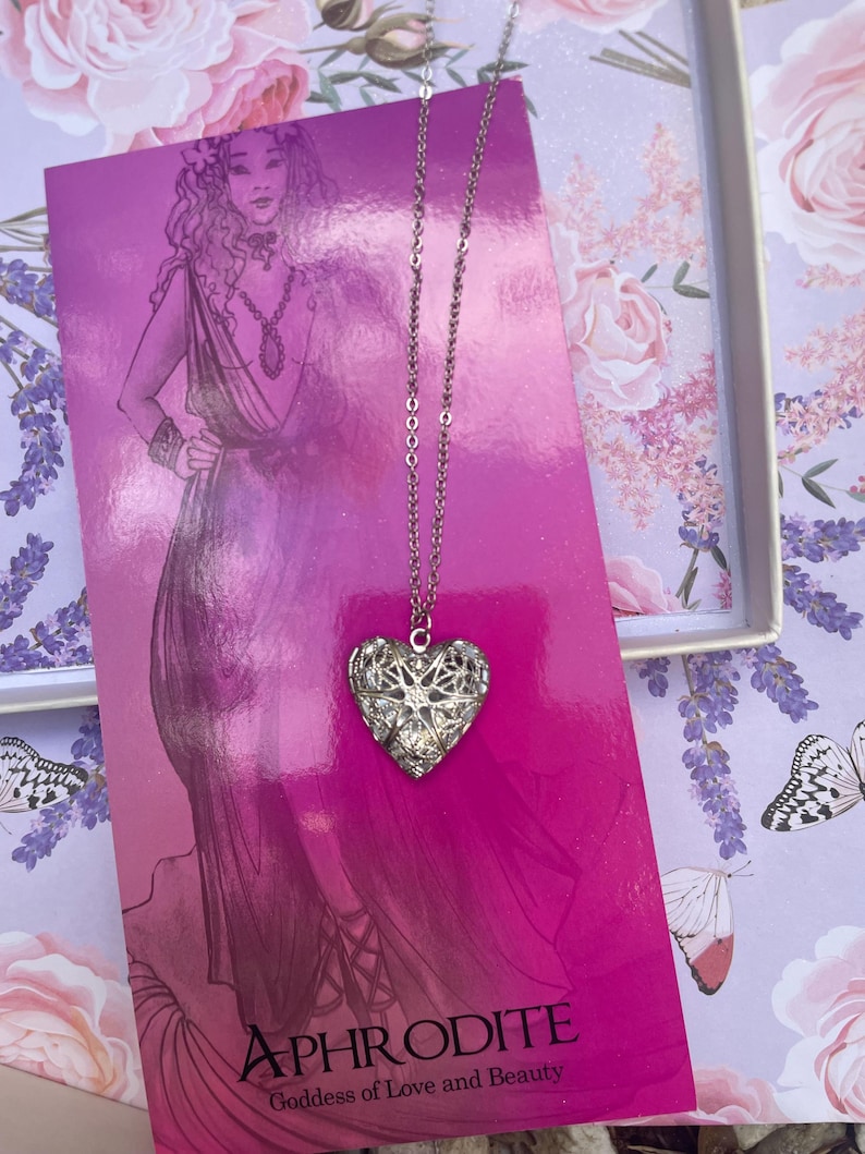 May include: A silver heart-shaped locket necklace with a delicate chain. The locket is adorned with intricate filigree designs and sparkling rhinestones. The necklace is displayed on a pink background with the text "APHRODITE Goddess of Love and Beauty".