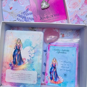 May include: A white box with a pink and blue floral design. Inside the box are several items, including a pink crystal, a card with a woman in a blue dress, and a card with the text "Goddess Aphrodite Affirmations". The text on the card reads "I am allowed to say no to others and "yes" to myself."