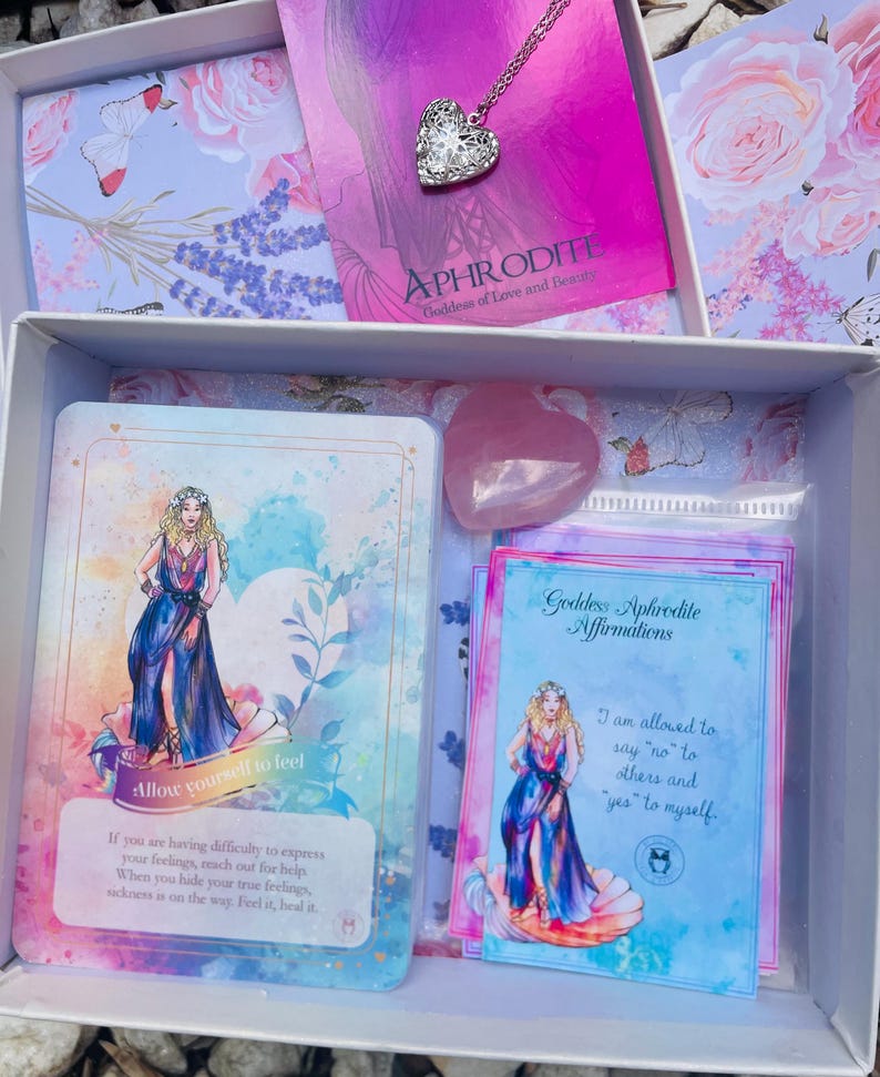 May include: A white box with a pink and blue floral design. Inside the box are several items, including a pink crystal, a card with a woman in a blue dress, and a card with the text "Goddess Aphrodite Affirmations". The text on the card reads "I am allowed to say no to others and "yes" to myself."