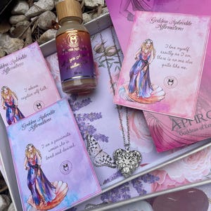 May include: A gift box set with a silver heart locket necklace, a bottle of self-love healing oil, and three affirmation cards featuring the goddess Aphrodite. The cards have pink and purple backgrounds with illustrations of the goddess and text that reads "Goddess Aphrodite Affirmations".