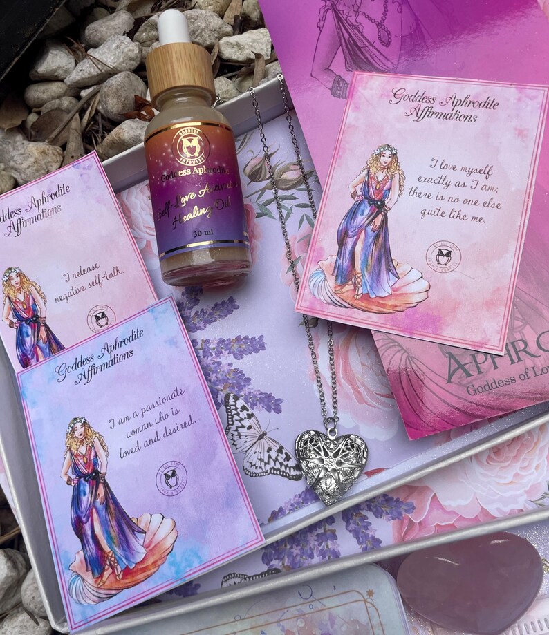 May include: A gift box set with a silver heart locket necklace, a bottle of self-love healing oil, and three affirmation cards featuring the goddess Aphrodite. The cards have pink and purple backgrounds with illustrations of the goddess and text that reads "Goddess Aphrodite Affirmations".