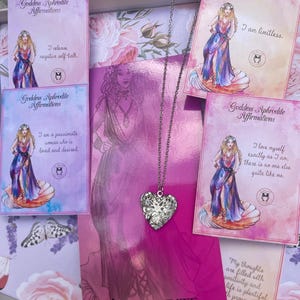 May include: A silver heart-shaped locket necklace with a delicate chain. The necklace is displayed in a box with four cards featuring illustrations of a woman and text about the Greek goddess Aphrodite. The cards include affirmations such as "I am limitless" and "I love myself exactly as I am."