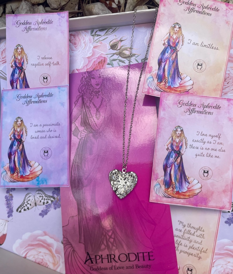 May include: A silver heart-shaped locket necklace with a delicate chain. The necklace is displayed in a box with four cards featuring illustrations of a woman and text about the Greek goddess Aphrodite. The cards include affirmations such as "I am limitless" and "I love myself exactly as I am."