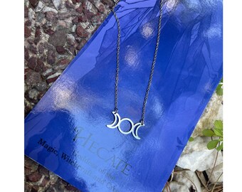 May include: A silver necklace with a triple moon pendant. The necklace is laying on a blue book with the text "Hecate Goddess of the Witchcraft and Crossroads" on the cover.