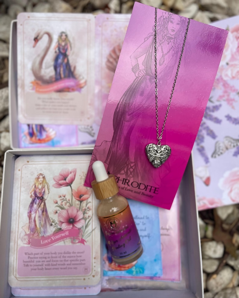 May include: A pink and purple box with a set of oracle cards, a bottle of essential oil, and a silver necklace with a heart pendant. The cards feature illustrations of a swan, flowers, and a woman. The bottle is labeled "Goddess Healing" and the necklace is labeled "Aphrodite Goddess of Love and Beauty".