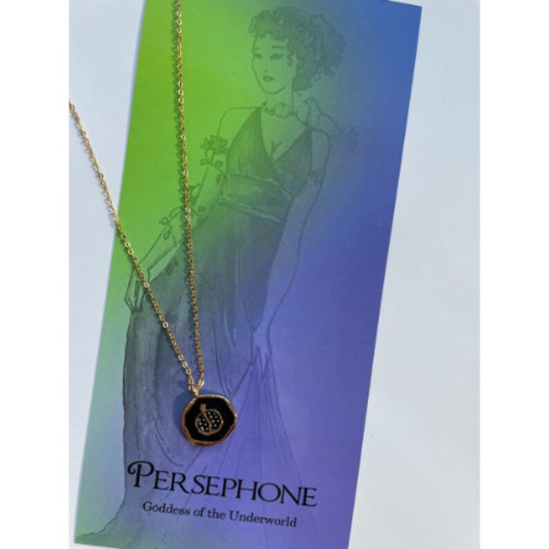 May include: Gold chain necklace with a black enamel pendant featuring a pomegranate design. The pendant has a gold rim and small clear stones. The necklace is displayed on a card with the text "PERSEPHONE Goddess of the Underworld".