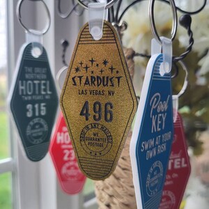 Puede incluir: Una colecci&oacute;n de llaveros de hotel de estilo vintage. Los llaveros tienen forma de llave de motel y presentan diferentes dise&ntilde;os y textos. Un llavero es dorado con el texto "STARDUST LAS VEGAS, NV" y el n&uacute;mero "496". Otro llavero es azul con el texto "Pool Key" y "SWIM AT YOUR OWN RISK".