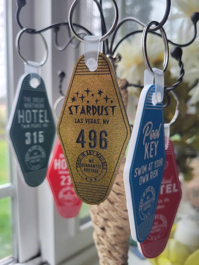 Puede incluir: Una colecci&oacute;n de llaveros de hotel de estilo vintage. Los llaveros tienen forma de llave de motel y presentan diferentes dise&ntilde;os y textos. Un llavero es dorado con el texto "STARDUST LAS VEGAS, NV" y el n&uacute;mero "496". Otro llavero es azul con el texto "Pool Key" y "SWIM AT YOUR OWN RISK".