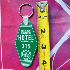 Puede incluir: Llavero de pl&aacute;stico verde con forma de llave de hotel con el texto "The Great Northern Hotel Twin Peaks, WA 315" y un peque&ntilde;o c&iacute;rculo con el texto "We Guarantee Postage".
