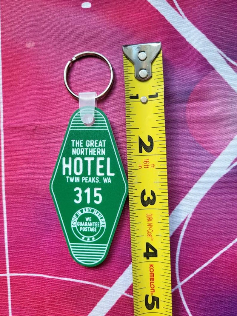 Puede incluir: Llavero de pl&aacute;stico verde con forma de llave de hotel con el texto "The Great Northern Hotel Twin Peaks, WA 315" y un peque&ntilde;o c&iacute;rculo con el texto "We Guarantee Postage".