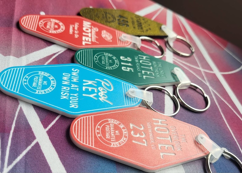 Puede incluir: Cinco llaveros de motel coloridos con diferentes dise&ntilde;os y textos. Los llaveros son rojos, verdes, azules y naranjas. El texto en los llaveros incluye "Pool Key", "Swim at your own risk", "Hotel", "Stardust Motel" y "The Overlook Hotel".