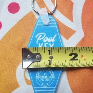 Puede incluir: Llavero azul de motel con el texto "Pool Key" en letras blancas cursivas. El llavero tiene un anillo de llave plateado y un lazo de pl&aacute;stico blanco. La parte posterior del llavero tiene un c&iacute;rculo blanco con el texto "We Guarantee Postage" y tres estrellas.