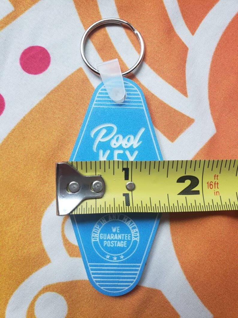 Puede incluir: Llavero azul de motel con el texto "Pool Key" en letras blancas cursivas. El llavero tiene un anillo de llave plateado y un lazo de pl&aacute;stico blanco. La parte posterior del llavero tiene un c&iacute;rculo blanco con el texto "We Guarantee Postage" y tres estrellas.