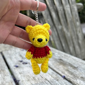 May include: A yellow and red crocheted bear keychain. The bear has a yellow body, a red shirt, and a black nose. The keychain is made of yarn and has a silver metal ring.