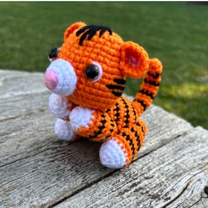 May include: A small crocheted tiger plush toy with orange and black stripes, white paws, and pink and black eyes.