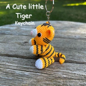 May include: A crocheted tiger keychain in orange and black stripes, with white accents on the paws and ears. The keychain has a silver ring and the text "A Cute little Tiger Keychain" in white letters.