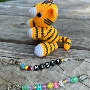 May include: A handmade orange and black crochet tiger toy with black eyes and white accents. Below are two keychains with the name "MISTY" spelled out in beads. The keychains have colorful beads and metal clasps.