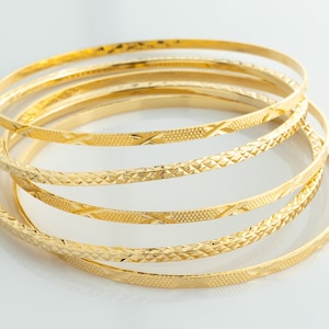 Puede incluir: Un conjunto de seis brazaletes de color dorado con un patrón de diamante. Los brazaletes están apilados uno encima del otro.