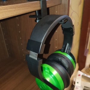 May include: A black and green headset is hanging on a shelf using a black clamp. The headset is attached to the clamp by the headband.