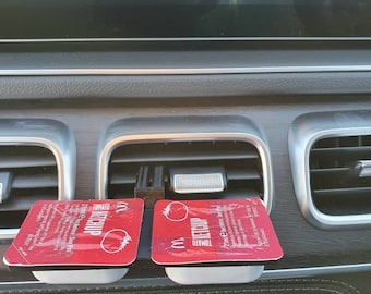 Puede incluir: Dos paquetes individuales de ketchup de tomate rojo y blanco se mantienen en un soporte de plástico negro fijado a la ventilación del tablero de instrumentos de un automóvil. El soporte tiene una base blanca. El texto en los paquetes dice "Ketchup de tomate" y "2 fl oz/59 ml".
