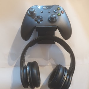 May include: A black Xbox controller and black headphones are mounted on a wall-mounted stand.