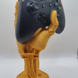 May include: A gold-coloured 3D printed hand holding a black Xbox controller. The hand is detailed with a mechanical design and is standing on a hexagonal base.