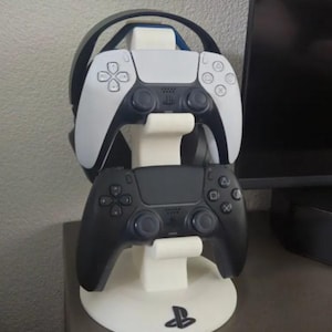 May include: A white PlayStation 5 controller and a black PlayStation 5 controller are displayed on a white stand with a PlayStation logo. The stand has a groove for a headset, which is not included.