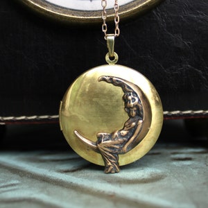 May include: A gold-toned locket pendant featuring a woman sitting on a crescent moon. The locket is on a gold chain.