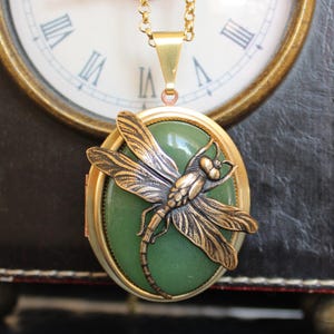 May include: A gold-toned necklace featuring a green oval pendant with a bronze dragonfly design. The dragonfly is detailed with intricate wing patterns. The necklace is displayed against a blurred background of a clock.
