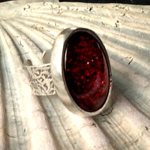 Puede incluir: Un anillo de plata con un centro ovalado lleno de purpurina rojo oscuro. La banda presenta un diseño calado decorativo. El anillo se encuentra sobre un fondo texturizado blanquecino, creando un contraste llamativo.