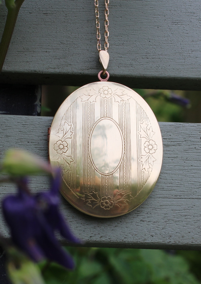 May include: A gold locket necklace with a detailed floral design and a blank oval space in the center. The locket is hanging from a chain on a gray wooden surface.